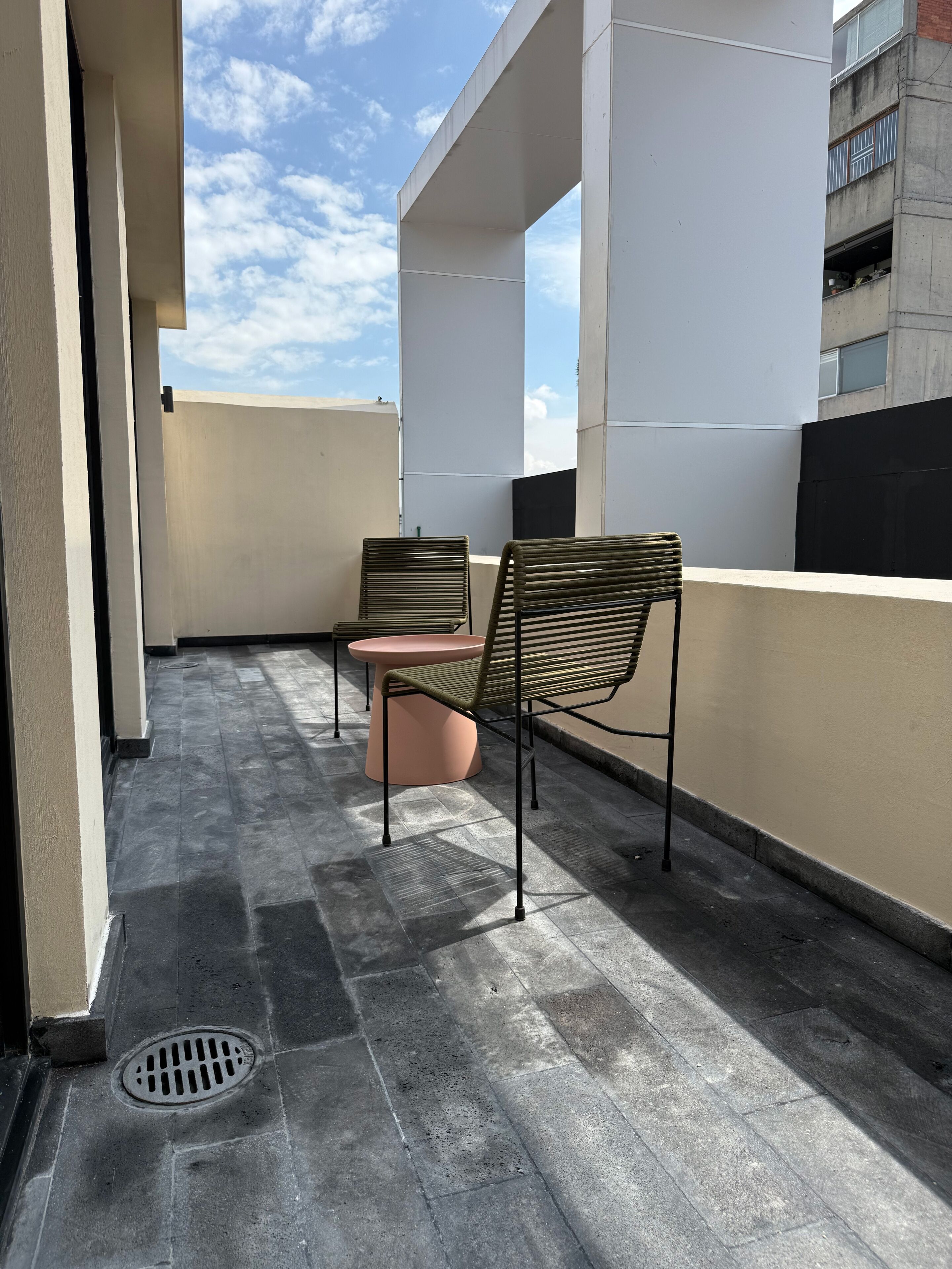 A rooftop terrace with two metal-framed chairs and a small round table, a vent on the floor, and a building in the background.