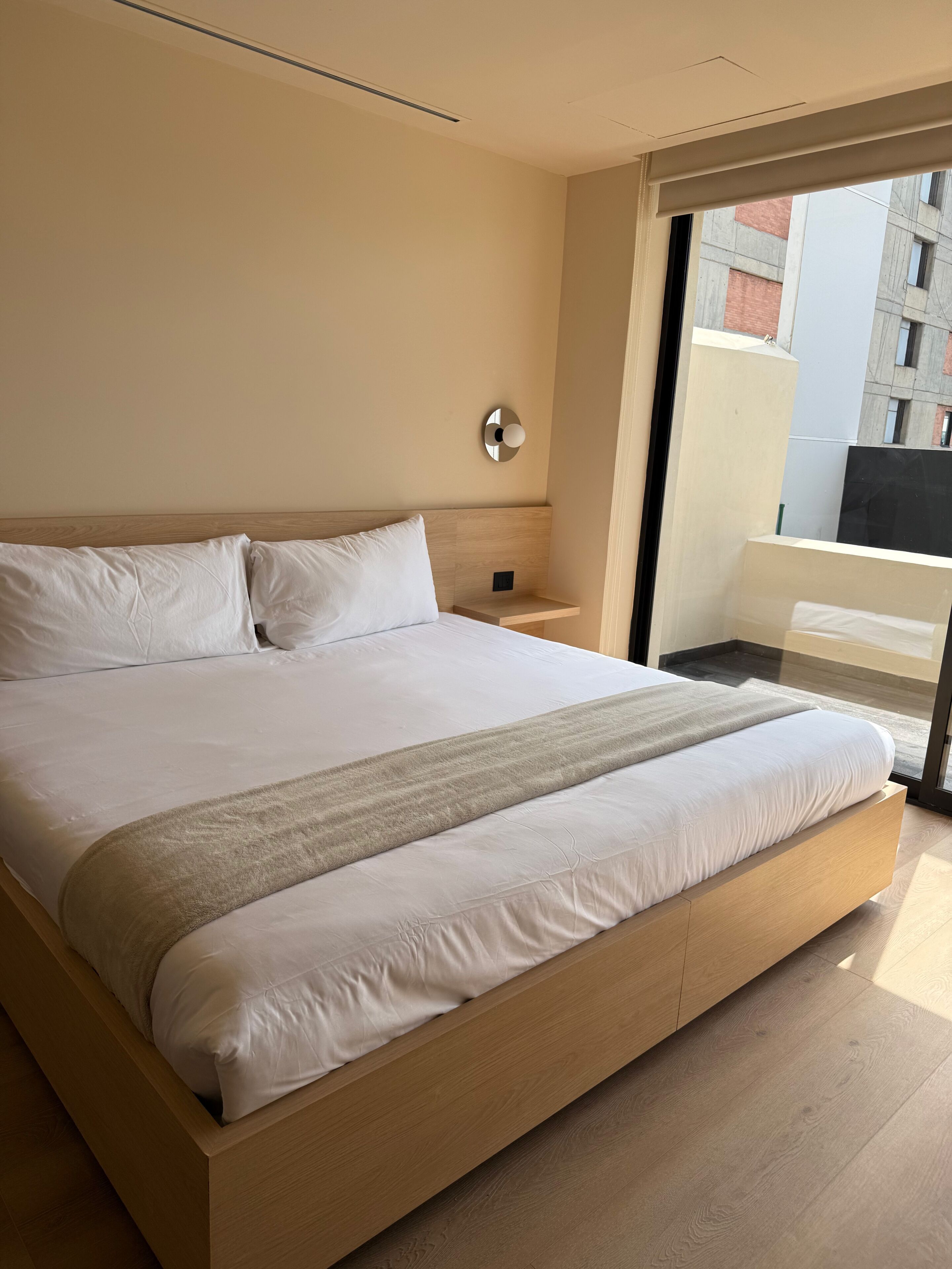 Ett modernt hotellrum med en stor säng, ett sängbord, en väggmonterad lampa och utsikt över staden genom ett stort fönster.