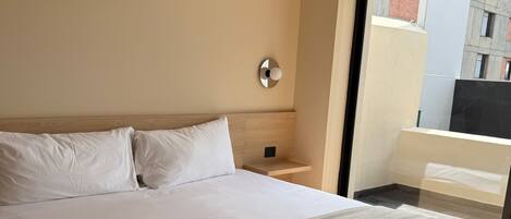 Ett modernt hotellrum med en stor säng, ett sängbord, en väggmonterad lampa och utsikt över staden genom ett stort fönster.