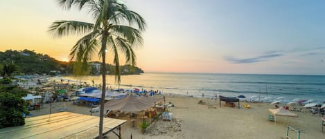 A beach with a palm tree, beach umbrellas, and a small building.