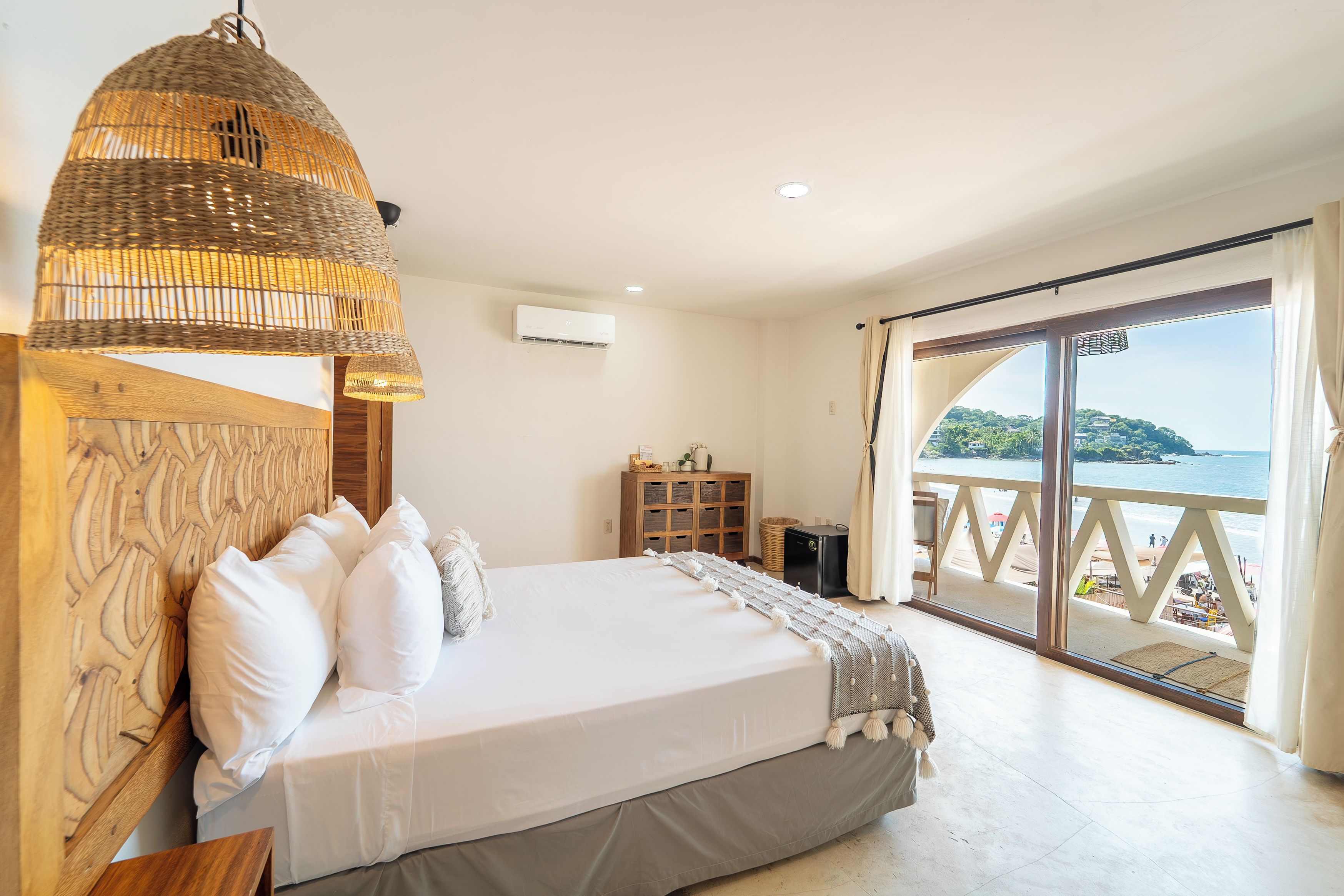 Un dormitorio con cama, una lámpara de mimbre, un armario de madera y vistas al mar.