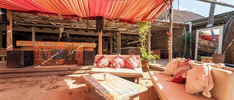 Un espace extérieur aménagé avec des sièges, un auvent rouge, du mobilier en bois et une scène équipée de haut-parleurs.