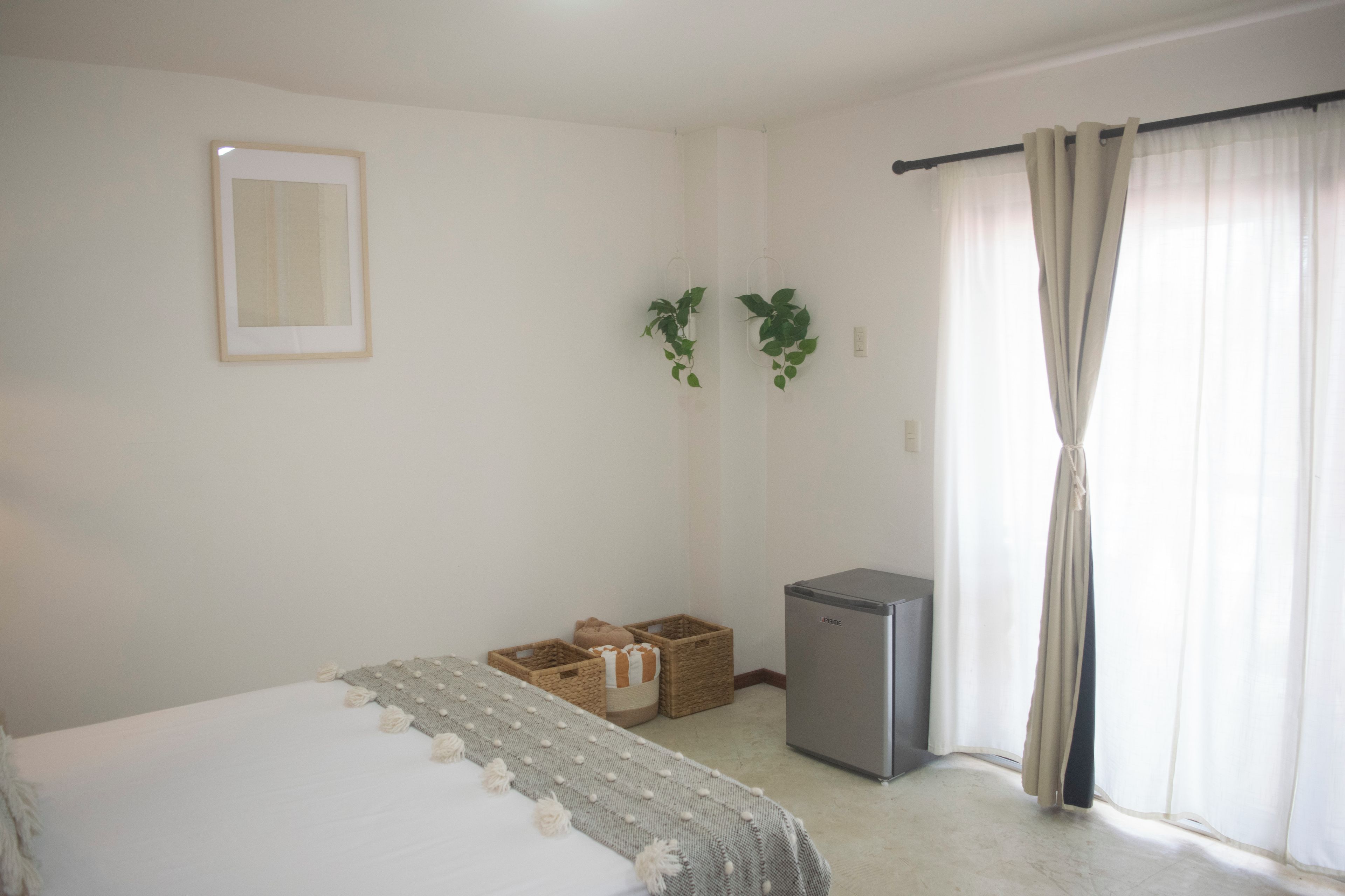 A bedroom with a bed, a small refrigerator, and a window with curtains.