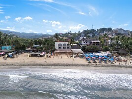 Una playa con una fila de sombrillas y sillas de playa, un edificio de techo rojo y palmeras al fondo.