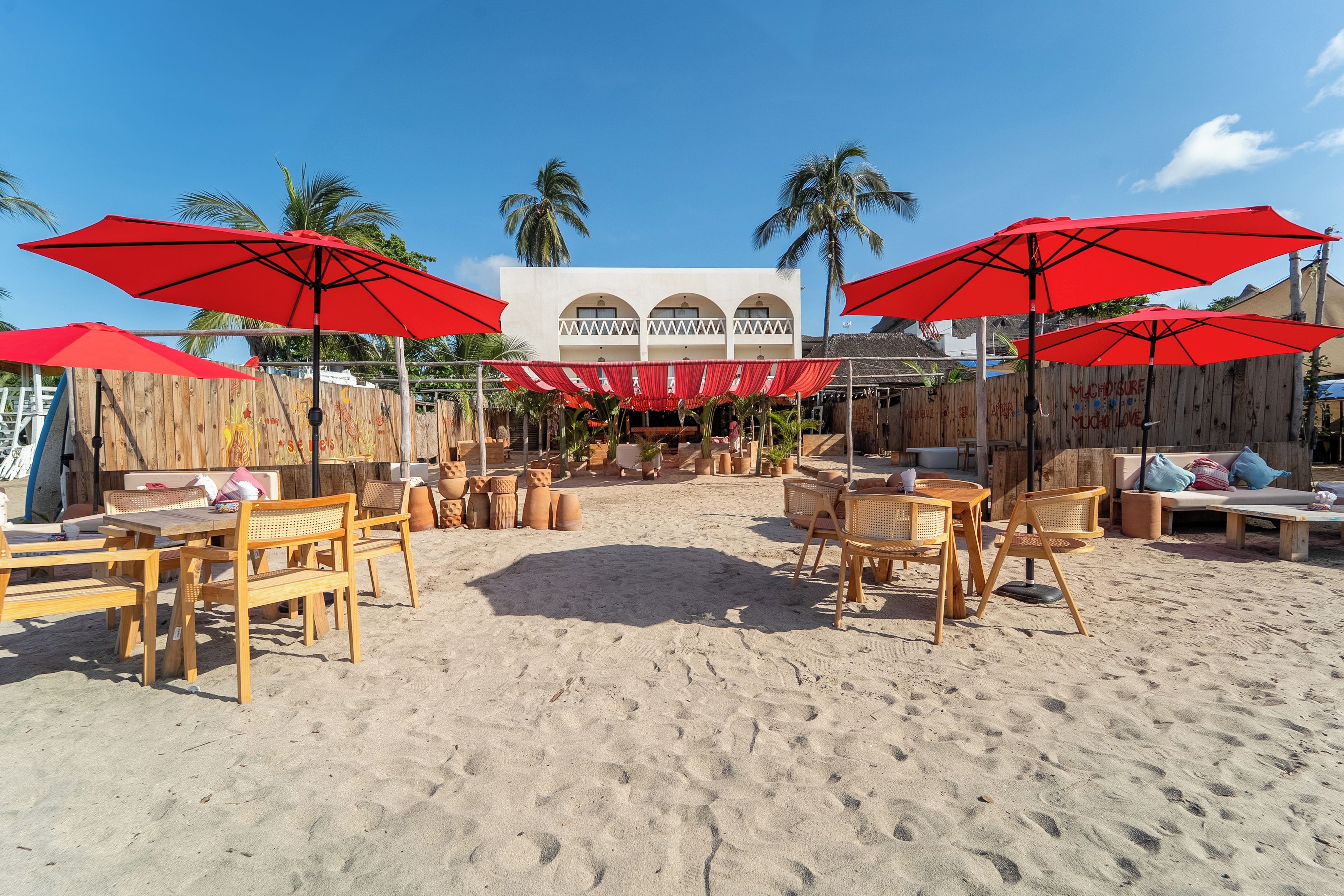 A beachside outdoor seating area with wooden furniture and red umbrellas.