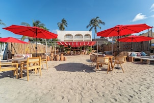 Un área de estar al aire libre frente a la playa, con mobiliario de madera y sombrillas rojas.