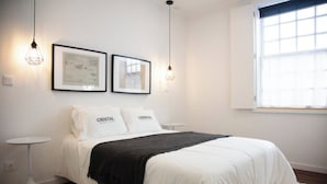 Ein ordentlich eingerichtetes Schlafzimmer mit einem Bett, zwei gerahmten Bildern, einem kleinen Nachttisch und einem Fenster mit Jalousien.