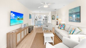 A spacious living room with a large flat-screen TV, a wooden console table, a white sofa, and a dining area with a glass-top table and chairs.