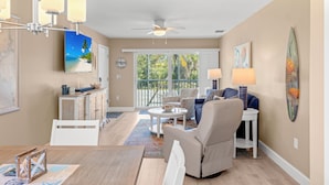 A spacious living room with a ceiling fan, a dining area with a table and chairs, and a view of a balcony with a door leading outside. - SeaShells Unit 40 – Island Getaway with Pool, Pickleball & Beach Access (Sanibel)