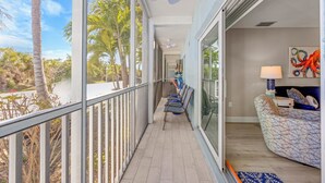 A balcony with a view of palm trees and a beach, a ceiling fan, and a seating area with a patterned sofa and a lamp. - SeaShells Unit 40 – Island Getaway with Pool, Pickleball & Beach Access (Sanibel)