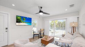 Una sala de estar con ventilador de techo, televisor en la pared, sofá, mesa de centro y vista al exterior a través de grandes ventanales.