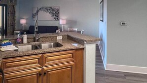 A compact kitchen with granite countertops, a stainless steel sink, wooden cabinets, and a small dining area with a table and chairs. - Shell Yeah! Oceanfront, Indoor Pool (Myrtle Beach)