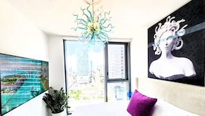 Un dormitorio moderno con una cama grande, una pintura de paisaje urbano, una escultura y vistas de edificios a través de un ventanal amplio.