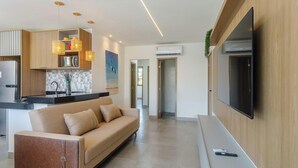 Una sala de estar moderna con sofá, televisor de pantalla plana montado en la pared y una cocina con armarios de madera e iluminación colgante.