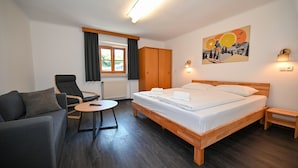 Une chambre d’hôtel moderne avec un lit en bois, un canapé, un fauteuil, une petite table et une fenêtre avec des rideaux.