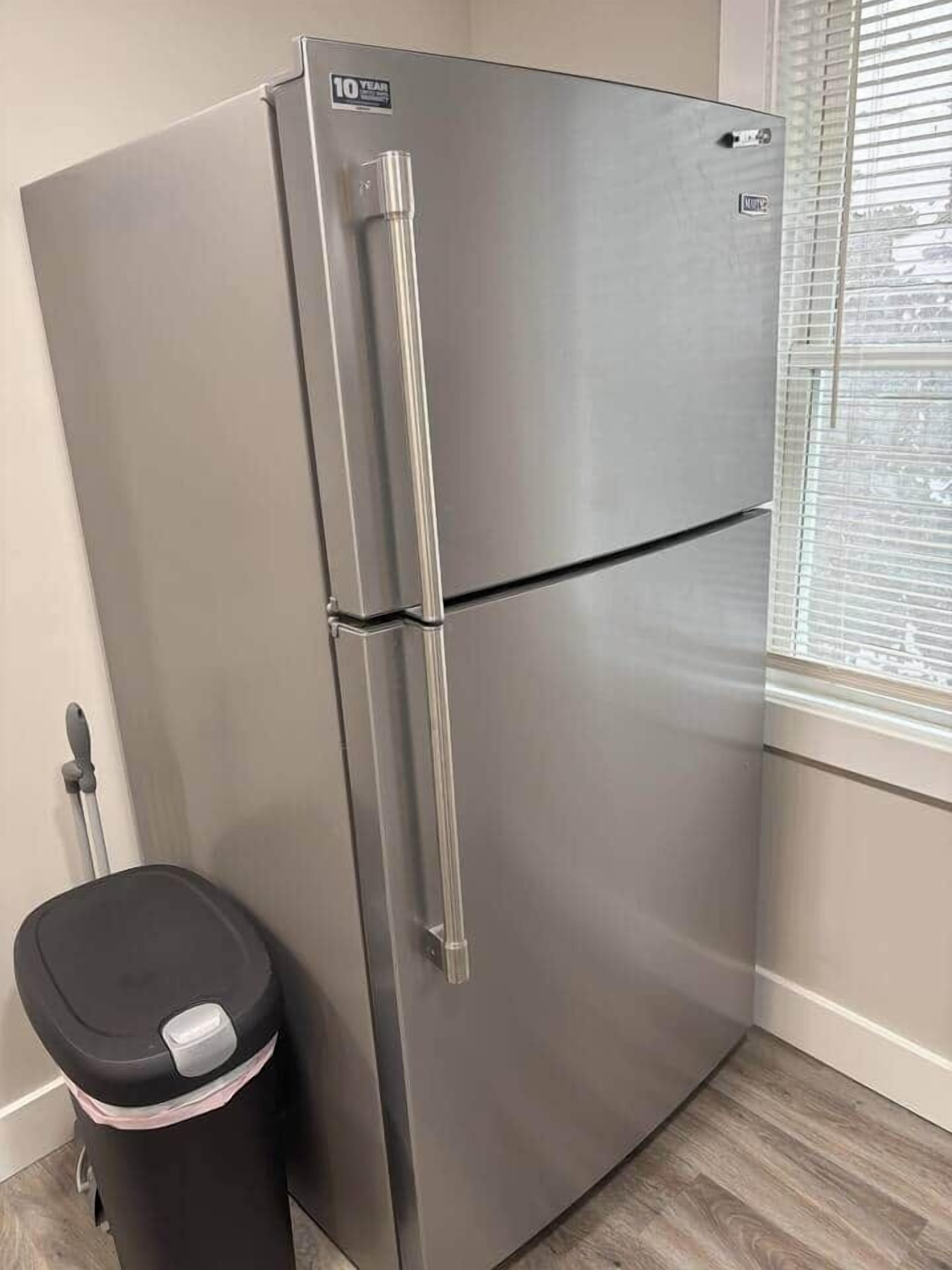 A stainless steel refrigerator with a 10-year warranty sticker, a black trash bin, and a window with blinds.