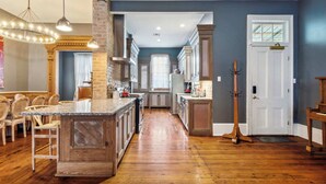 A spacious kitchen with wooden flooring, a central island, and a dining area with chairs. - Victorian Villa w Heated Pool & Hot Tub 5BR|6BA (New Orleans)