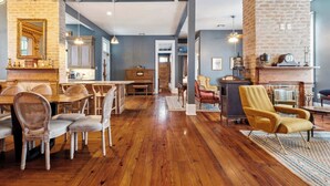 A spacious living area with a wooden floor, a dining table with chairs, a yellow armchair, and a fireplace with decorative items. - Victorian Villa w Heated Pool & Hot Tub 5BR|6BA (New Orleans)