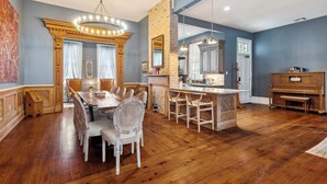 A spacious dining area with a wooden table and chairs, a chandelier, and a kitchen area with a large island and hanging lights. - Victorian Villa w Heated Pool & Hot Tub 5BR|6BA (New Orleans)