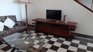 Una sala de estar con un sofá, una mesa de centro de vidrio, un televisor sobre un mueble de madera y una lámpara.