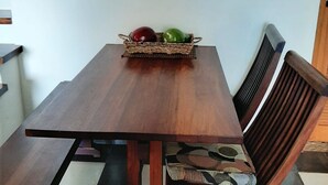 Una mesa de comedor con una cesta tejida llena de fruta, una silla con asiento de mimbre y un adorno colgado en la pared.