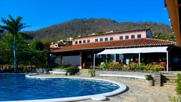Un área de piscina con un edificio al fondo, rodeada de vegetación y un paisaje montañoso.