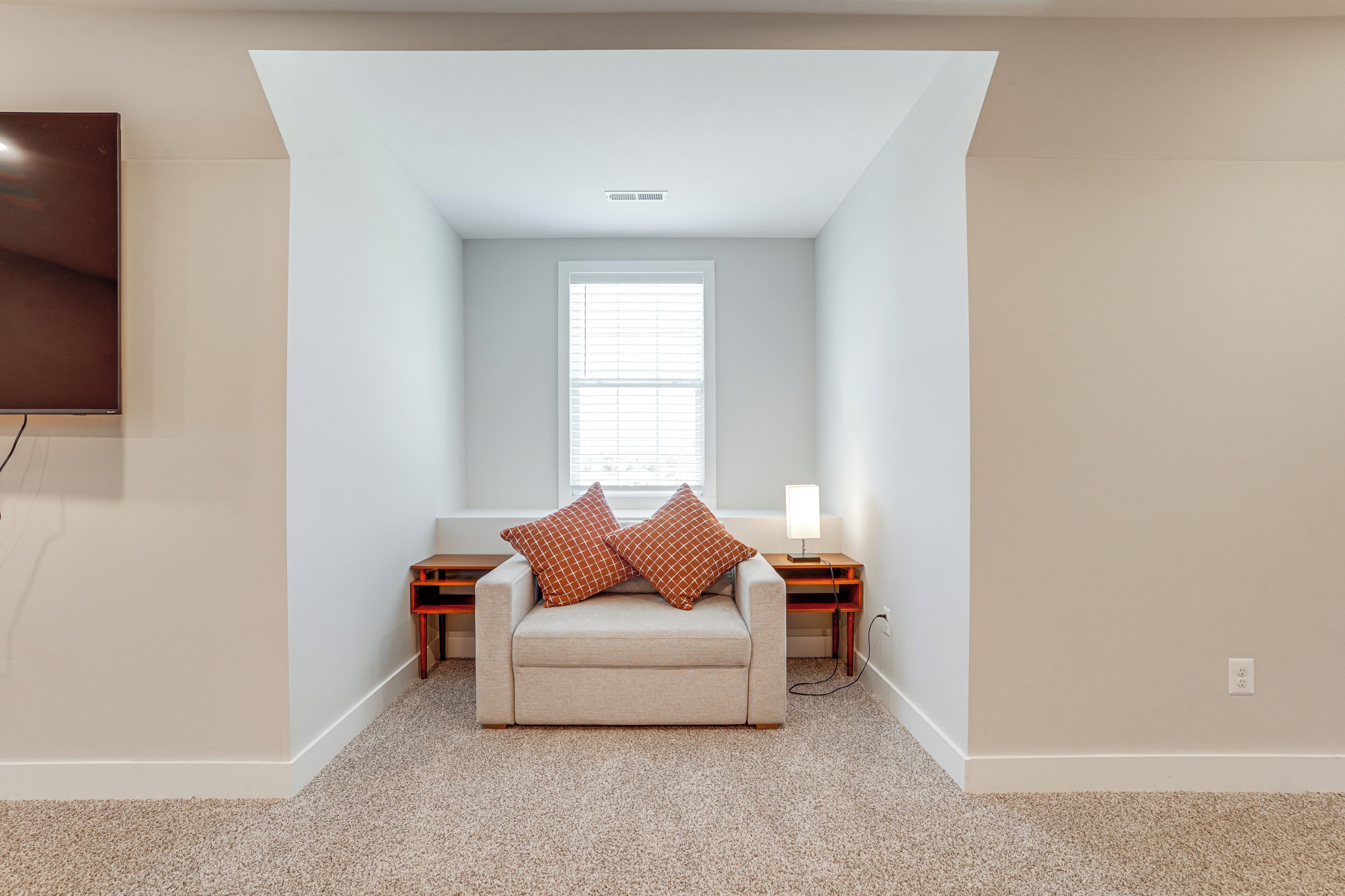 A small, cozy room with a beige sofa, two pillows, a side table with a lamp, and a window with blinds.