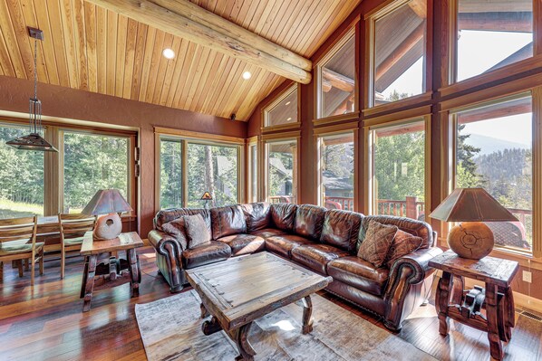 A spacious living room with a large leather sectional sofa, wooden coffee table, and large windows offering a view of the outdoors. - Ski-In/Out, Hot Tub: Spacious Retreat in Whitefish (Whitefish)