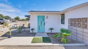 Una casa moderna con una puerta turquesa, un pequeño patio con sillas y plantas de palma.