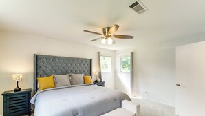Una camera da letto con testiera trapuntata, un letto con copripiumino grigio e bianco, una panca ai piedi del letto, un comodino con una lampada e una ventola a soffitto.