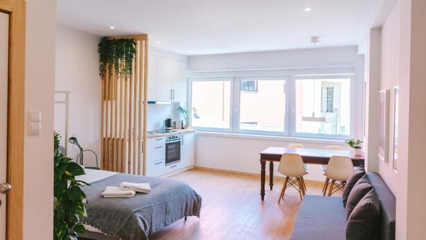 A modern, well-lit room with a bed, a dining table with chairs, a kitchenette, and a large window offering a view of the outside. - Porto serenity studio (União de Freguesias do Centro)