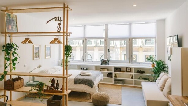 A modern bedroom with a bed, a wooden shelf unit, a window with a view, and a white sofa.