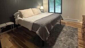 Un dormitorio con cama, mesita de noche, lámpara, ventilador de techo y vistas a árboles a través de una ventana.