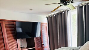 Quarto com cama, televisão sobre um armário de madeira, ventilador de teto e janela com cortinas.
