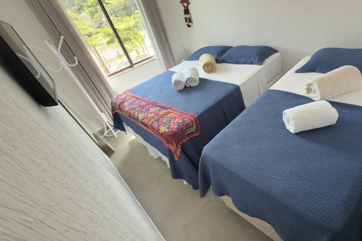 A hotel room with two beds, each with a blue bedspread and white pillows. There is a window with a view of greenery outside.
