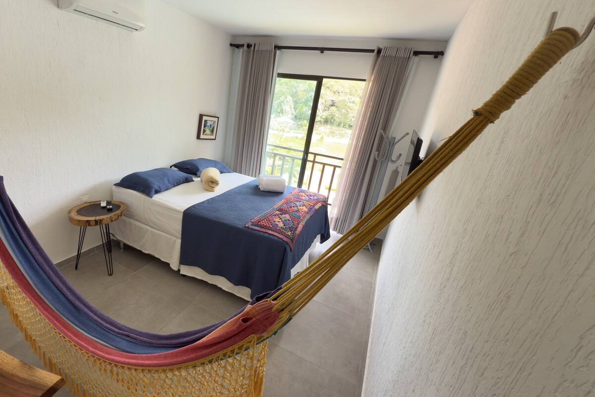 A bedroom with a bed, a small table, a hammock, and a balcony with curtains.