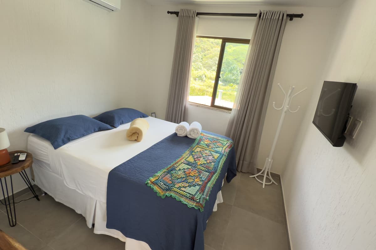A neatly made bed with a blue blanket and pillows, a bedside table with a lamp and remote, a window with curtains, and a coat rack.