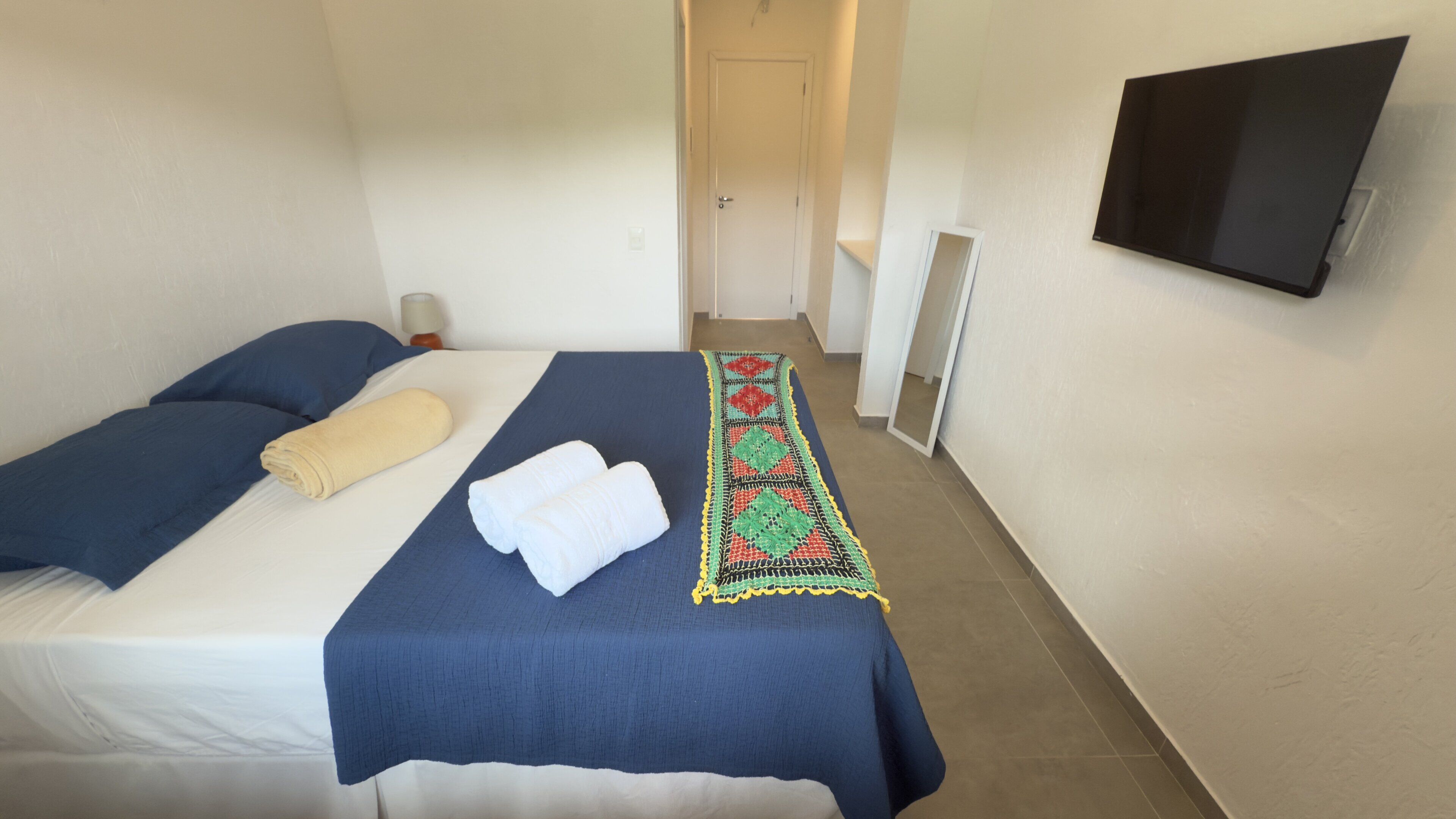 Una habitación de hotel con una cama, un televisor montado en la pared y un pasillo que conduce a otra habitación.