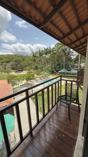Una terraza de madera con vista a una piscina, palmeras y un autobús al fondo.