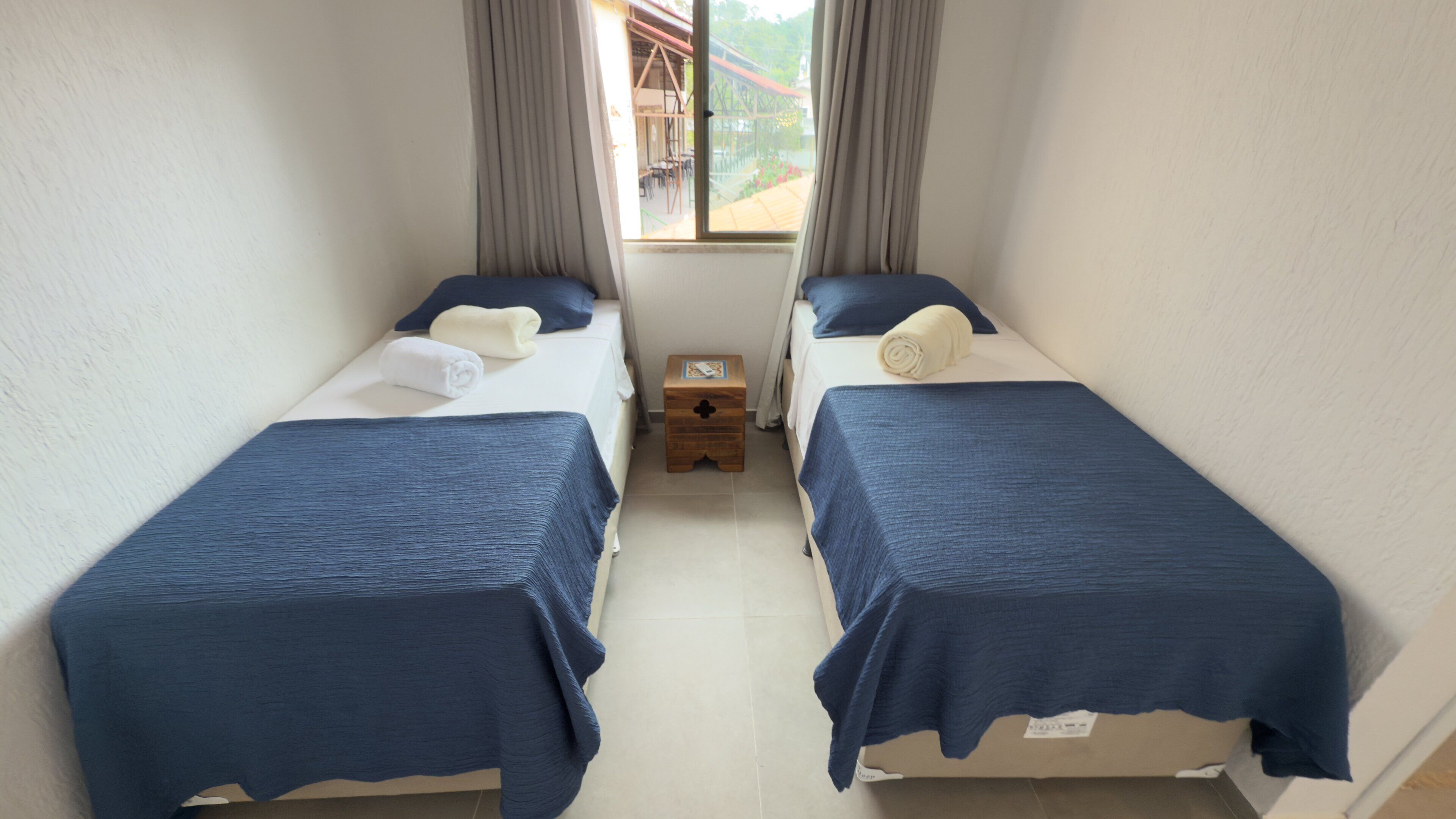 Et værelse med to senge, hver med et blåt sengetæppe og puder, et trænatbord og et vindue med gardiner.