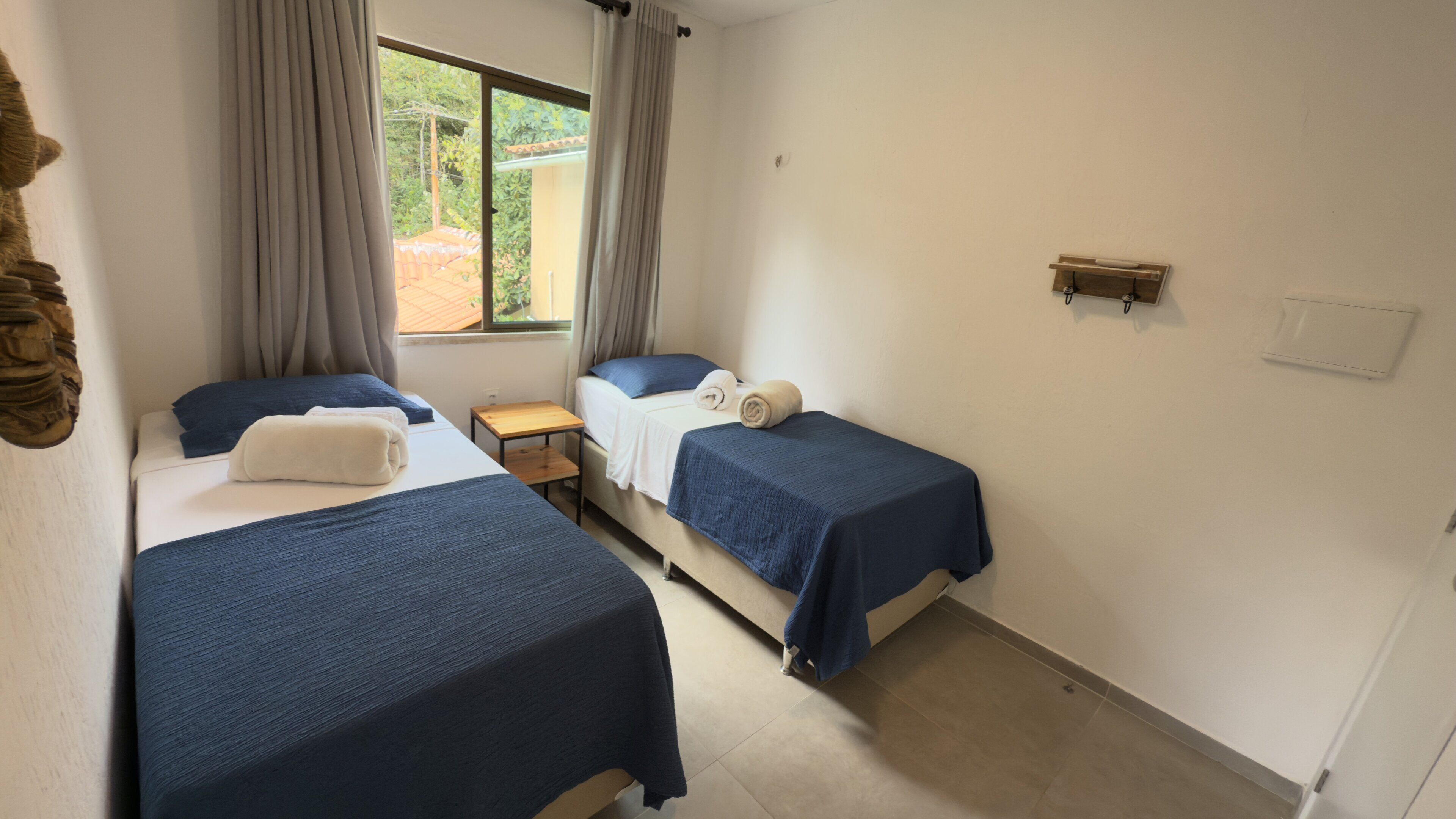 A room with two beds, each with a blue bedspread and pillows, a small wooden table, and a window with curtains.