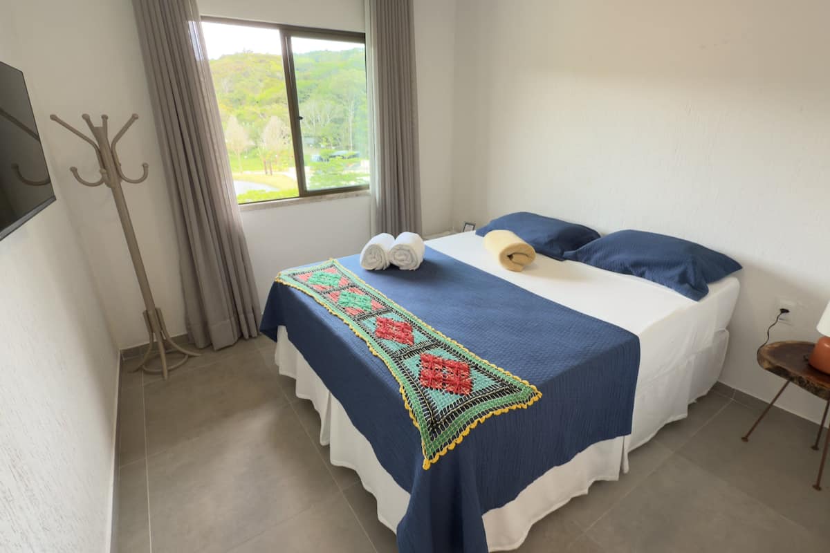 A bedroom with a bed, a TV mounted on the wall, a window with curtains, and a small table with a lamp.
