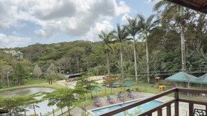 Una terraza de madera con vista a una piscina, palmeras y un parque infantil.