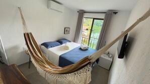 Una habitación con cama, una hamaca, una mesita, una mesilla de noche y un ventanal con cortinas.