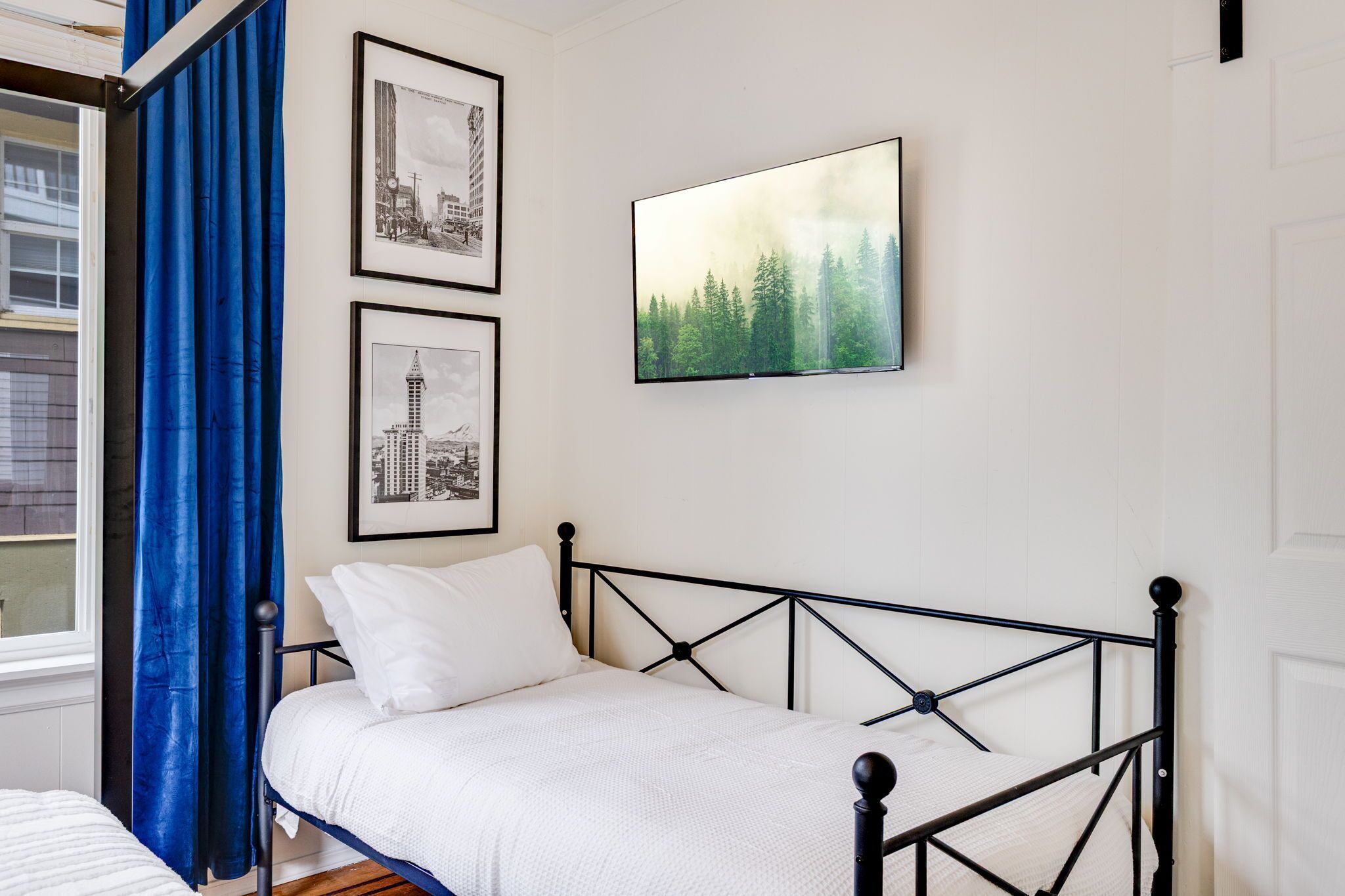 Een slaapkamer met een bed, fotolijstjes en een groot raam met blauwe gordijnen.