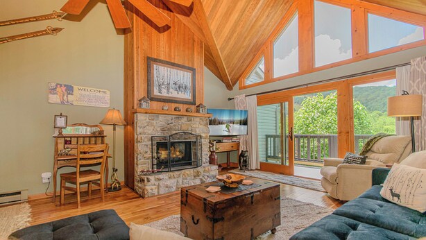 A spacious living room with a stone fireplace, wooden ceiling beams, and large windows offering a view of the outdoors. - SlopeView Chalet:Steps 2 Slopes, Fire Pit, Huge Deck, AC, Pet Friendly, Sleeps 9 (Beech Mountain)