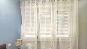 A room with two windows covered by sheer white curtains, a bedside lamp, and a small table with a decorative item.