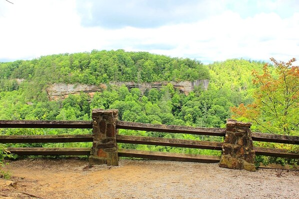 A wooden fence with stone posts, a gravel path, and a view of a forested hill with a cliff.