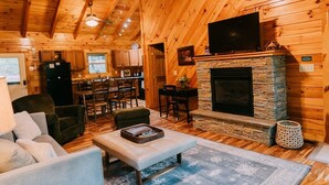 A cozy cabin interior with a stone fireplace, wooden walls, a dining area, and a living room with a sofa and chairs. - Cabin, Ensuite Bedrooms, Hot Tub, Firepit, Woods (Campton)
