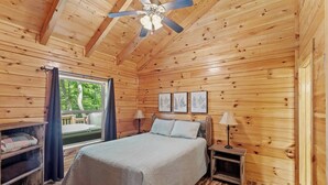 A wooden cabin bedroom with a bed, two bedside tables, a ceiling fan, and a window with a view of greenery. - Cabin, Ensuite Bedrooms, Hot Tub, Firepit, Woods (Campton)